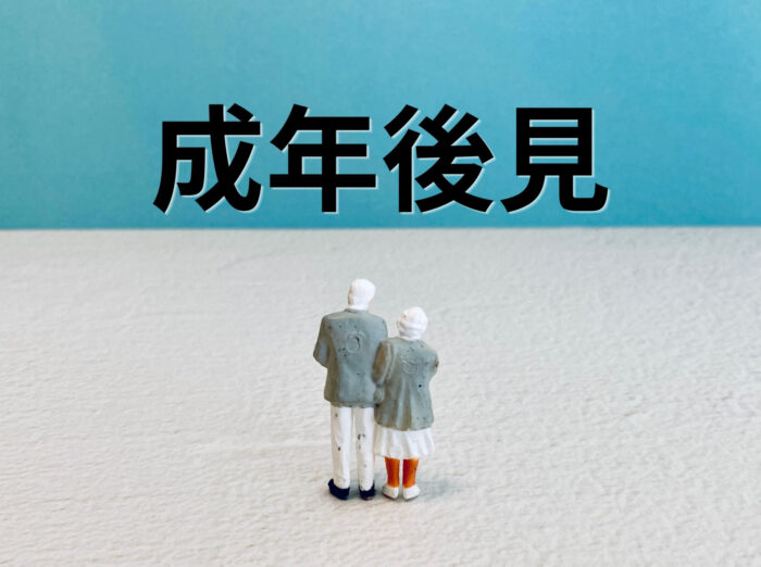 「成年後見」の文字の前に並んで立つ高齢夫婦のミニチュア人形の後ろ姿